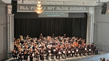 View from balcony of an orchestra on a stage. In front of those musicians are a few singers with stands and music. At the top of the stage area are the words, All Passes Art Alone Endures.