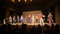 Over a dozen actors of various demographics walk about a theater stage. They are all dressed in monochromatic, nondescript outfits.