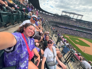 Selfie at a baseball game taken by Charlotte looking out at the crowd seated along the first baseline. Charlotte is wearing a purple Hawaiian shirt, and all looking at the camera are smiling.