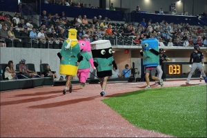 A group of four costumed characters race each other down a baseball field sideline. Yellow, which Evan is within, appears to be in the lead.