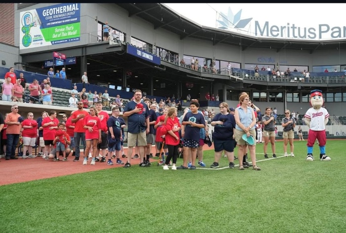 A crowd on the baseball field as the national anthem is sung. Evan is in the mascot costume!