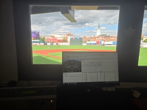 View of a desktop computer setup in what appears to be a control booth over a baseball field, big windows behind the desktop displaying the field, home plate obscured by the computer monitor.