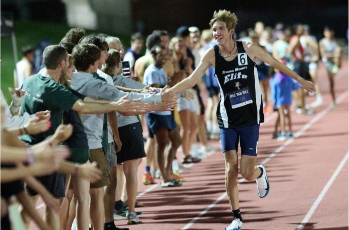 Isaac runs down a track lane toward the camera at an angle, his arm outstretched to high five the crowd along the sidelines.