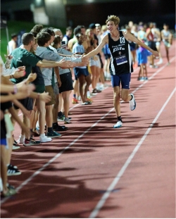 Isaac runs down a track at an angle toward the camera, his right arm outstretched to high five members of the crowd along the sidelines who are cheering for him.