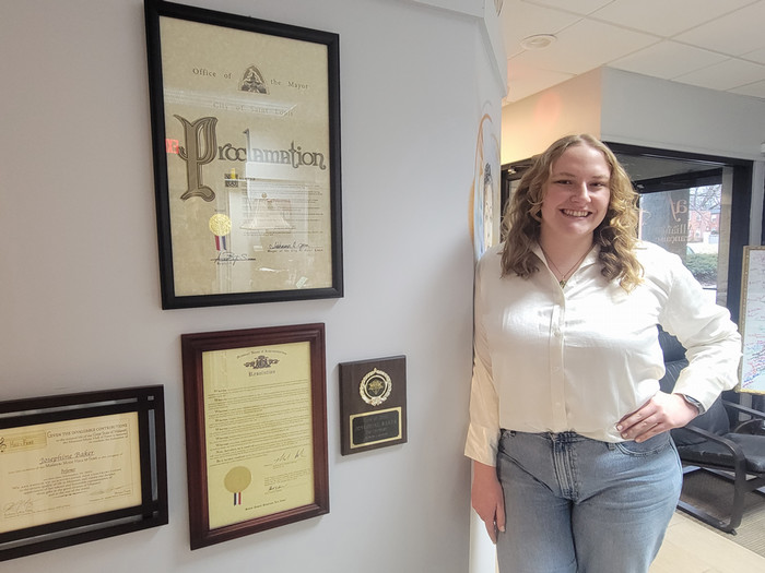 Katie standing next to a wall of displayed papers in the office of the Alliance Française, including a proclamation from the Mayor of Saint Louis