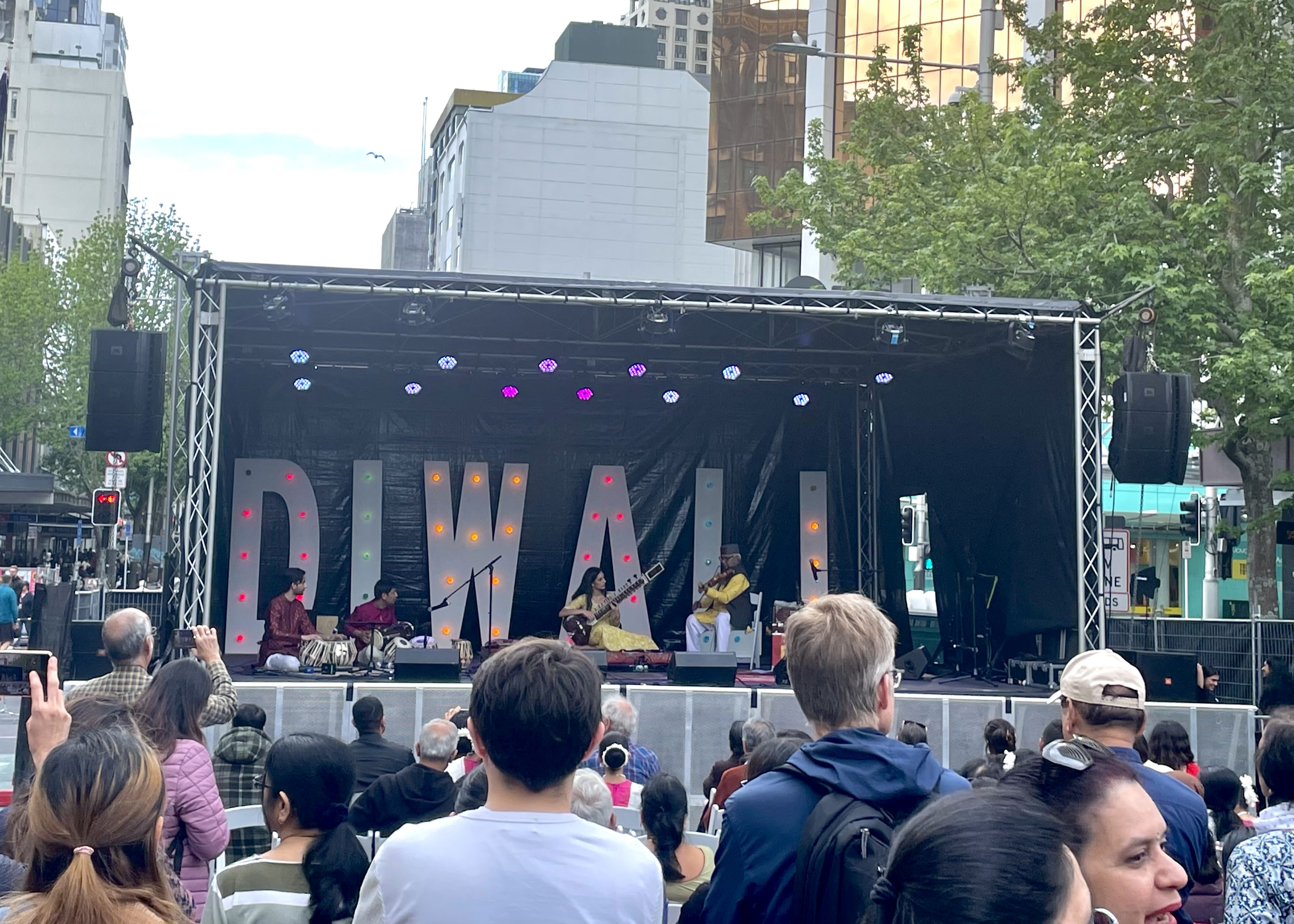 A band, sitting in front of a large sign reading "DIWALI", in front of a crowd