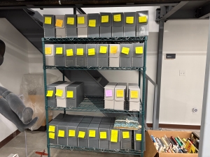 A shelving unit holds four rows of grey boxes, most of which have yellow sticky notes on them