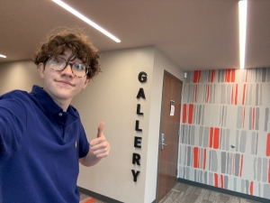 Selfie of Orion in a blue polo flashing a thumbs up, a sign for the gallery he's in visible on the wall to his left