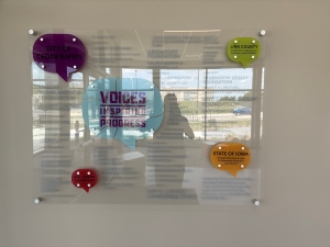 Clear, reflective plaque with a few colorful splashes in the corners with individual words on them. In the reflection, you can see the figure taking the photo, Orion.