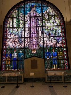 Beautiful, multi-colored stained glass window. The top three-fourths depict a woman in a flowing lavender dress looking down to the bottom fourth which contains multiple people looking up to her