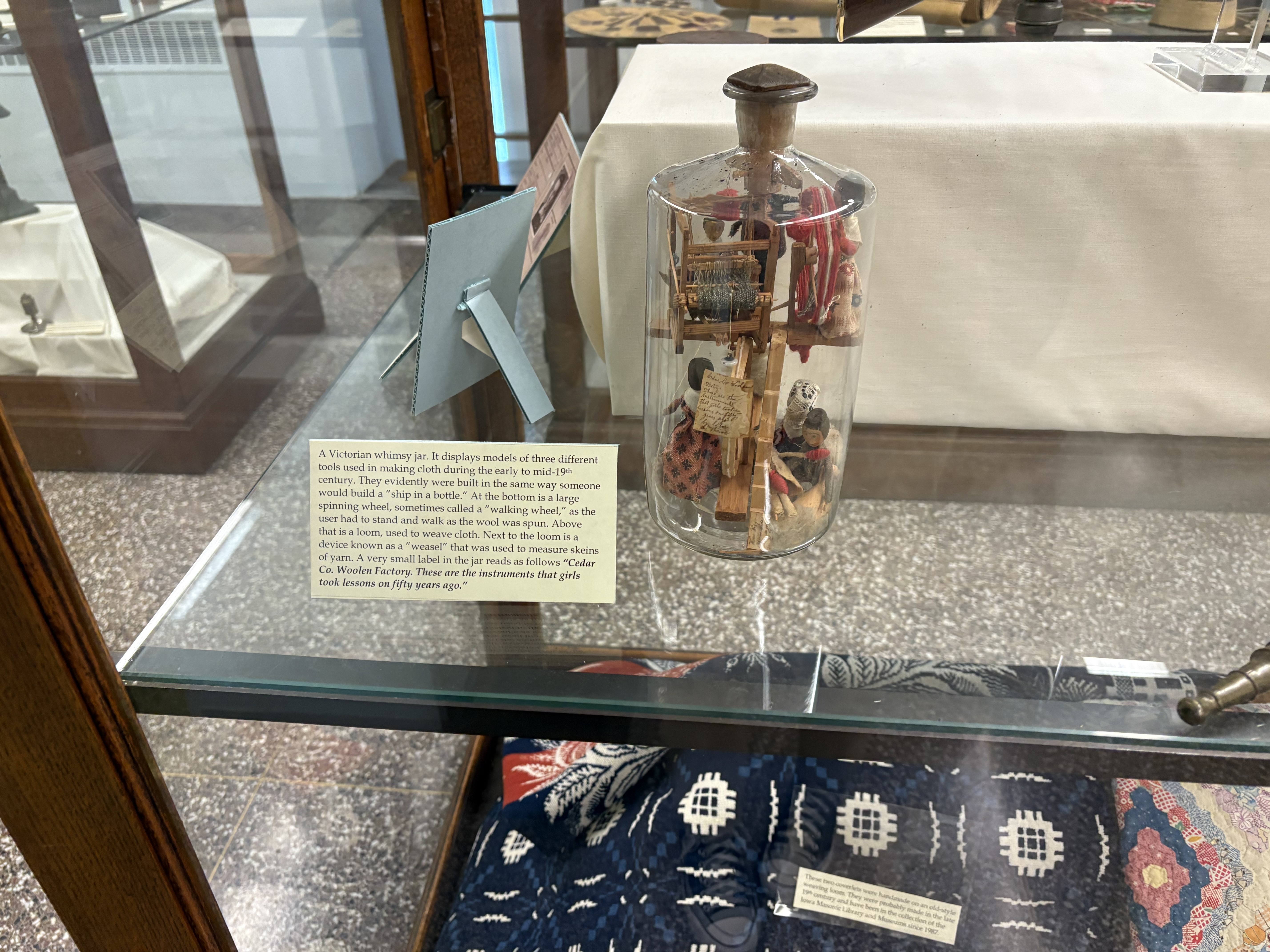 Display of a see-through glass jar with various objects inside it sitting on a see-through glass display case with a quilt visible beneath it
