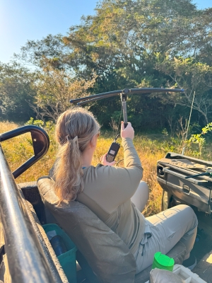 Peyton faces away from the camera with a tool that looks like a crossbow combined with a pickaxe in her right hand facing out into a tree-lined grassy area.