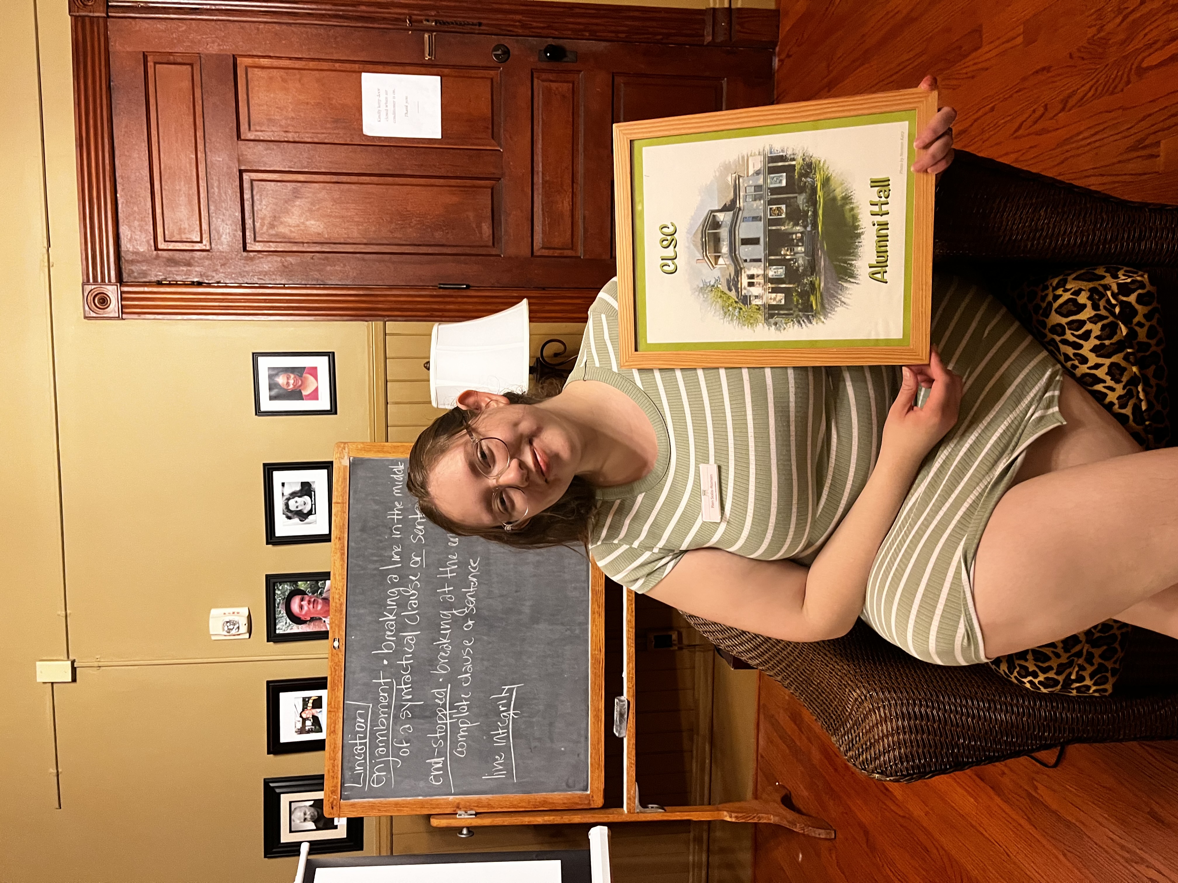 Photo of Ren holding a framed artistic depiction of the CLSC Alumni hall. Ren is sitting in a chair in what is called the Reading Room