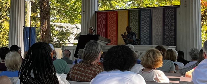 View of two speakers on a stage in an outdoor enclosed ampitheatre-style space from the viewpoint of the audience looking from the right at the stage