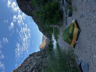 First camping spot in the Gunnison Gorge along the river on 4th of July weekend. There is a large American flag and two small ones near a pitched tent along the shore of a waterbed gorge.