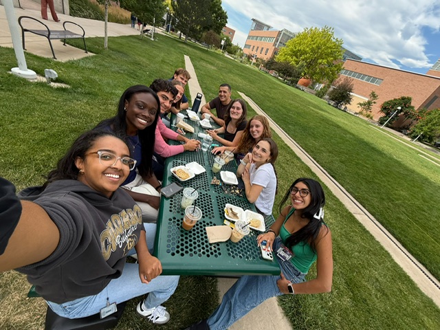Group of about six to eight people of various races gathered together at a picnic table looking at the camera (the camera seeing the table vertically)