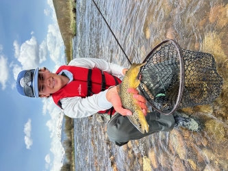 Sean in fishing gear holding a brown trout in his right hand and smiling at the camera