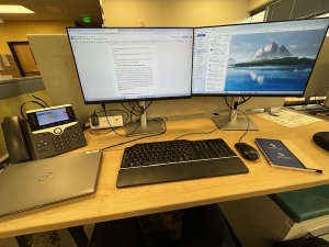 Sean's daily workstation setup for writing the manuscript at the hospital. It is a desk with two large monitors, a keyboard and mouse, a phone, and a closed laptop to the left.