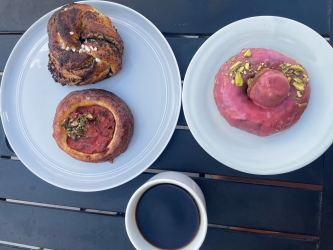 The image shows two white plates and a white coffee mug on a wooden picnic table. The white plate at the top of the image has a gallete of some kind and a bagel that hasn't yet been sliced. The plate below has what appears to be a glazed donut.