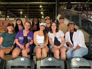 Group photo of Sophie and 8 other interns. Sophie is on the left edge in the front row with four others in her row and four in the back. All are smiling, and some are wearing baseball clothing.