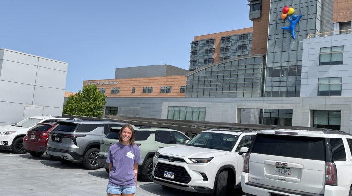 Sophie in a purple shirt and denim shorts standing in front of the Children's Hospital Colorado building, the sign and logo visible on the building behind her.