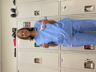 Sophie poses in her blue scrubs in a locker room setting. She does not yet have her scrub cap or mask on, so we can see her smiling face.