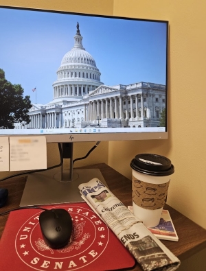 Photo shows a computer desk with one monitor visible with the U.S. Capitol building as the computer's desktop background. A rolled up copy of a The Quad-City Times newspaper and a to-go coffee cup sit to the right of the mouse.