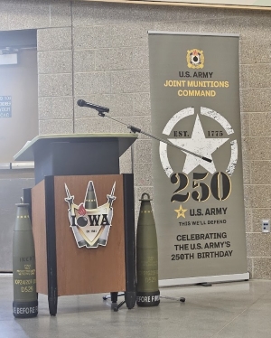 Image shows a podium with military weapons surrounding a design of the word Iowa. Next to the podium is a banner celebrating the 250th birthday of the Army