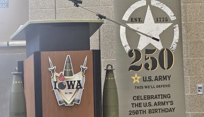 Image shows a podium with military weapons surrounding a design of the word Iowa. Next to the podium is a banner celebrating the 250th birthday of the Army