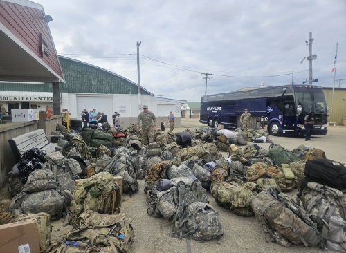 A sea of duffel bags with camouflaged design sit in a parking lot near a long-distance transit bus.