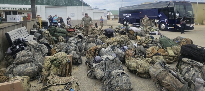 A sea of duffel bags with camouflage design sit in a parking lot next to a long-distance transit bus. 