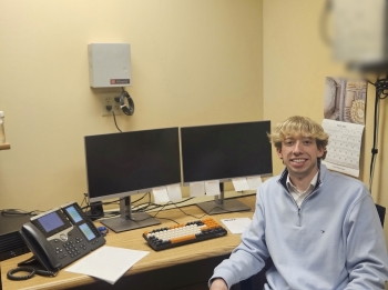 Tyler is seated at his computer desk, his back facing a wall with a calendar hanging on it to the side of the desk. Two computer monitors, turned off, sit on the desk as well as a keyboard, mouse, and phone unit.