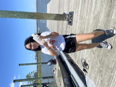 A girl holding a clear bottle of water on a boat dock, surrounded by water. The girl has a white shirt, black and white shorts, and block and white converses on. 