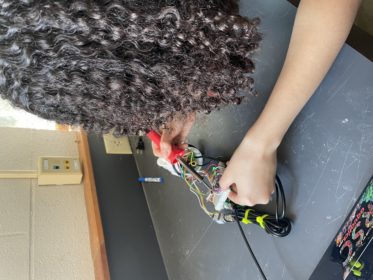 A girl with curly hair (face not visible) holding a red soldering iron to breadboard. 